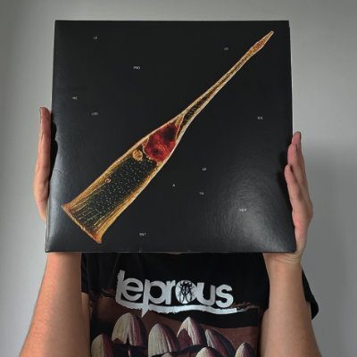Młoda kobieta trzyma w ręce płytę zespołu Leprous Melodies Of Atonement. Nie widać jej twarzy. Zasłania ją płyta winylowa. Okładka płyty przedstawia zdjęcie planktonu w kształcie dzwonka na czarnym tle.