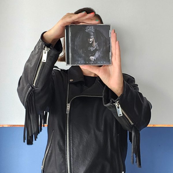 Kobieta w czarnej, skórzanej kurtce z frędzlami trzyma w dłoniach okładkę płyty cd Ozzy'ego Osbourne'a Ordinary Man. Płyta zasłania jej twarz.